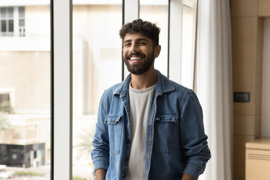 Successful and satisfied. Cheerful candid millennial male in stylish youth closing stand near window at modern office. Happy young student enjoy good day at university. Smiling employee feel motivated