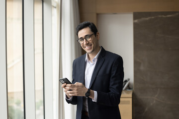 Fototapeta premium In step with time. Portrait smiling successful modern day businessman executive ceo stand by window hold smartphone gadget look at camera. Young manager in glasses use phone for business communication