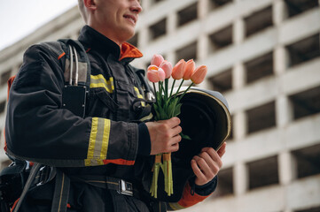 Present of flowers in hands. Professional firefighter is standing outdoors against big unfinished...