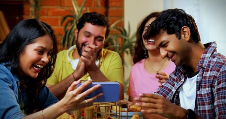 Watching funny video in cafe, Indian Asian young college friends or university students laughing loudly while eating samosa, sipping hot chai, enjoying comedy clip on smartphone indoors joyfully