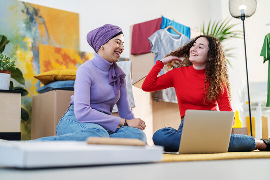 Two young women sit on the floor surrounded by moving boxes and clothes. They smile and look at each other while using a laptop, sharing a happy moment.