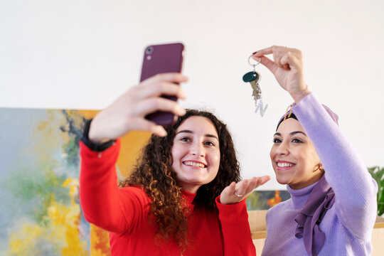 Two diverse young women are taking a selfie indoors. One holds up keys, celebrating a new home or successful purchase together. They are happy and sharing their joy.