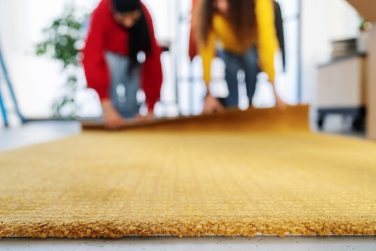 Two people are installing a new yellow rug on the floor. They are working together on a home renovation project. The focus is on the texture of the carpet.