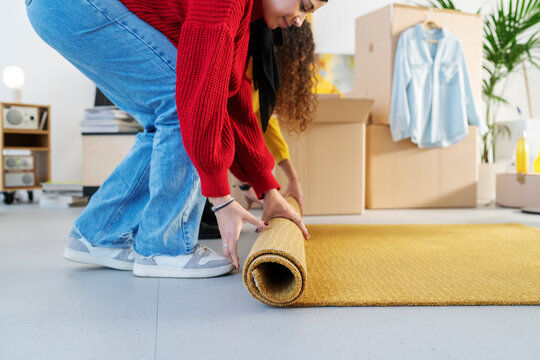Two young women are unrolling a yellow rug on the floor of their new home. Moving boxes are in the background. They are settling into their new place.