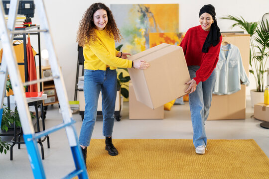 Two young women are carrying a large cardboard box together. They are smiling and working as a team in a room with moving boxes, clothes, and furniture, suggesting a move or organization.