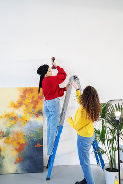 Two women are working together on a home renovation project. One woman uses a drill while standing on a ladder, and the other woman assists her from below.