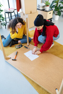 Two young women are assembling a piece of furniture on the floor. They are following instructions and using tools like a screwdriver and hammer. Moving boxes are nearby.