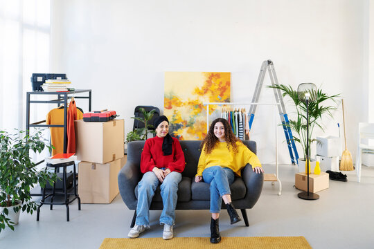 Two young women, one in a hijab, sit on a sofa in a room filled with moving boxes and items. They smile, taking a break from setting up their new space.