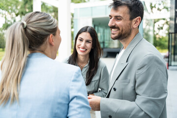 Diverse business team of corporate professionals in formal attire collaborating outdoors, engaged in strategic discussion while analyzing data on laptop in front of modern glass office building.