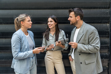Diverse business team of corporate professionals in formal attire collaborating outdoors, engaged in strategic discussion while analyzing data on laptop in front of modern glass office building.