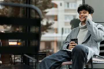 Young man talking on his cell phone while sitting in a chair with a coffee cup in his hand in front of a window