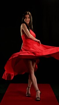 Stunning Black-Haired Woman in Ruby Red Dress and High Heels Gracefully Posing Against a Black Background, Captured in Super Slow Motion. Vertical Aspect Ratio.