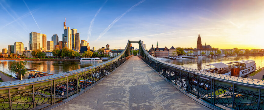 Panoramic view of the old iron bridge Eiserner Steg crossing the Main river in Frankfurt, Germany, with the skyline behind during summer sunrise