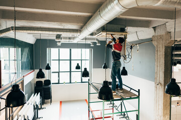 Construction worker performing maintenance on ceiling equipment in modern workspace during daylight hours