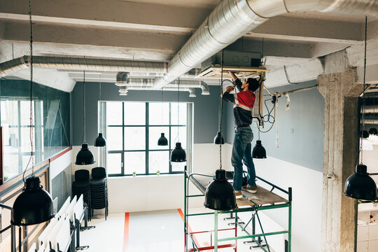 Construction worker performing maintenance on ceiling equipment in modern workspace during daylight hours