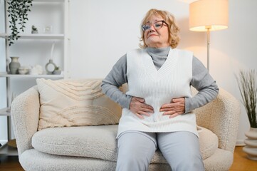 Mature woman suffering from stomach ache sitting on a bed in the bedroom. Senior woman sitting on...