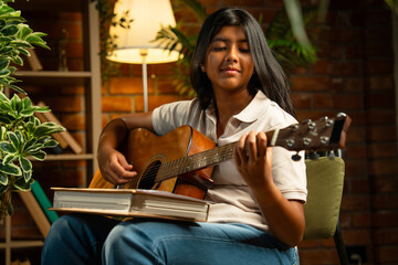 Indian teenage girl strumming guitar and writing songs in dimly lit cozy room against brick wall