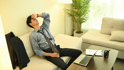 A Burnout businessman in a loosened tie rests on the sofa, struggling to stay awake as he continues...