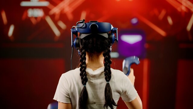 The little girl use tracking controllers in her hands, moves actively immersing herself in modern game. The child wearing VR headset plays game. Behind girl there is screen with blur background image