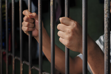 Hands of a prisoner behind prison bars on black background