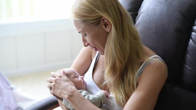 Mother-blonde in white T-shirt sits on armchair and holds sleeper baby in olive overalls on hands. Newborn - happiness for family. New worries and lifestyle.