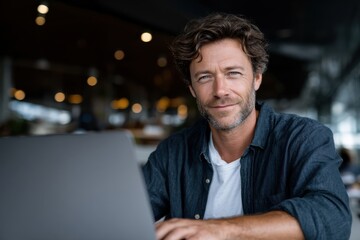 A confident man leans forward at his laptop, sporting a friendly smile that exudes professionalism in a modern, stylish workspace filled with natural light.