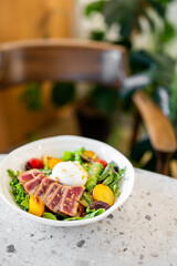 A gourmet salad bowl with seared tuna, poached egg, cherry tomatoes, olives, and mixed greens, served on a textured countertop. The blurred background features a cozy dining setup with wooden elements