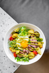 Fresh salad with mixed greens, cherry tomatoes, roasted potatoes, sliced beef, and a poached egg with green dressing, served in a white bowl on a marble countertop.