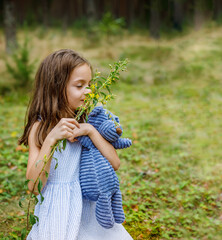 Little girl standing in a peaceful forest, gently holding a blue teddy bear while smelling a delicate flower