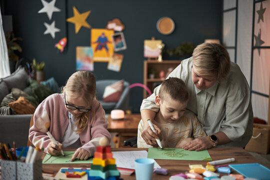 Woman assisting two young children with an art and craft project at home in a cozy living room filled with art supplies and colorful decorations, fostering creativity and learning