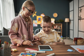 Girl playing together with boy with disability in tablet device in cozy living room, showing visible excitement and enjoyment while exploring screen content