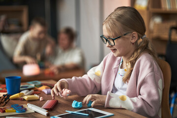 Young girl with glasses engaging in creative play with colorful clay at a table filled with toys...