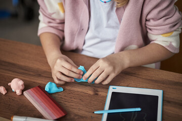 Child sculpting playful clay figures on wooden table with digital gadget nearby, focusing on...