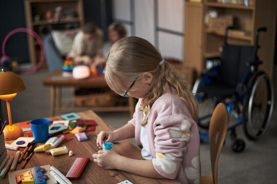 Young girl focusing on creating an art project at a desk while teacher and another student engaging in background in classroom setting providing supportive environment