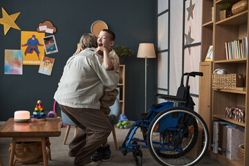 Woman gently hugging young boy with disability in wheelchair in a cozy living room decorated with...