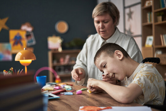 Young boy with disability engaging in creative activity with help from teacher in classroom setting. Teacher supervising and assisting student with crafts at wooden table - Powered by Adobe