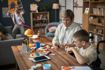 Mother and son with disability engaged in making crafts at wooden table, surrounded by art supplies...