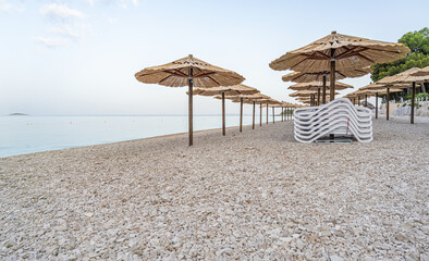 A serene pebble beach lined with rustic straw parasols under a blue sky beside calm sea waters.