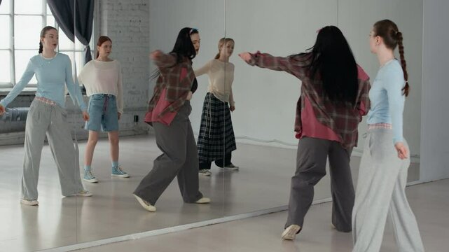 Long shot of four Caucasian female zoomers in streetwear practicing hip hop moves by mirror wall in dance studio
