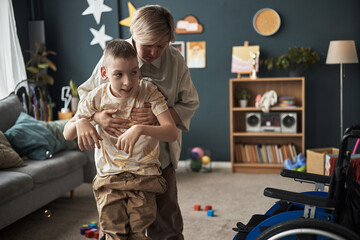Caregiver assisting child with disability for physical exercise in a cozy living room filled with...