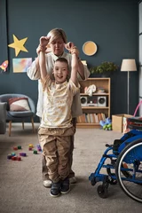 Vitrage gordijnen Child with disability practicing standing and mobility with help from an adult caregiver. Colorful room filled with toys and wheelchair in foreground © DragonImages