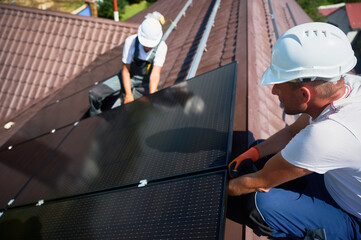 Workers building solar panel system on rooftop of house. Two men installers in helmets installing photovoltaic solar module outdoors. Alternative, green and renewable energy generation concept.
