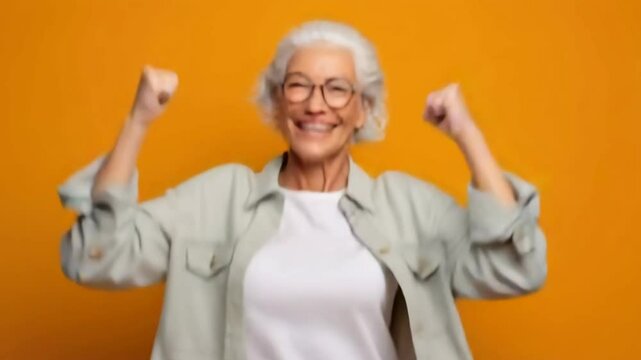Joyful elderly woman celebrating with raised fists on a vibrant yellow background, expressing happiness and positivity in her golden years