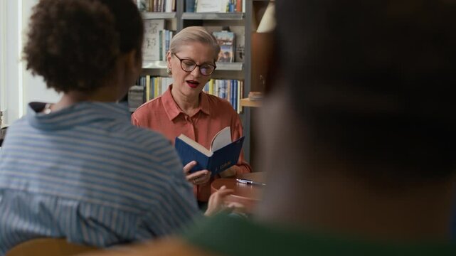 Smiling mid-aged Caucasian female author introducing her latest book to small group of listeners during cozy book event, surrounded by literature and welcoming bookstore ambiance