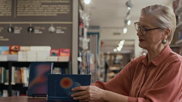 Side-view waist up of confident mid-aged Caucasian female author signing multiple book copies handed to her by bookstore event attendees during literary gathering