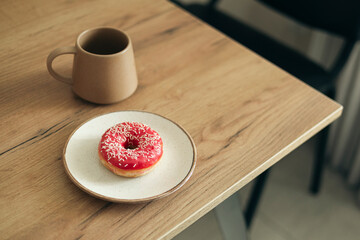 Pink donut and a cup on a table, capturing cozy breakfast and sweet treat.