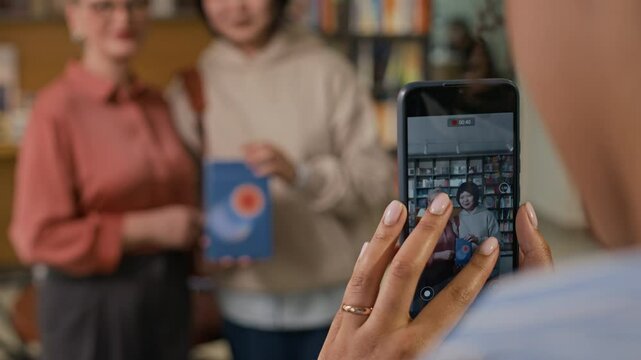 Over-the-shoulder shot of fan taking video of famous author and reader posing with her book during bookstore event, capturing joyful moment through smartphone screen