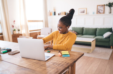 Young woman in yellow sweater typing on laptop and smiling, listening through wired earphones while working at home, surrounded by colorful notes and stationery supplies