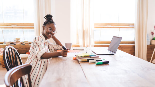 African American woman sitting at wooden desk, smiling and creating in notebook, surrounded by books and digital tools in bright creative home studio