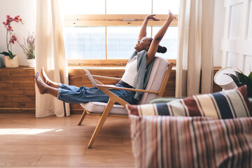 Young African woman leaning back with hands behind head and smiling while resting from laptop work at home, embracing calm, joyful freelance lifestyle in modern cozy room